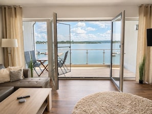 A modern living room with a sofa, coffee table, and chairs, a balcony with a view of the water and boats, and a TV on the wall.