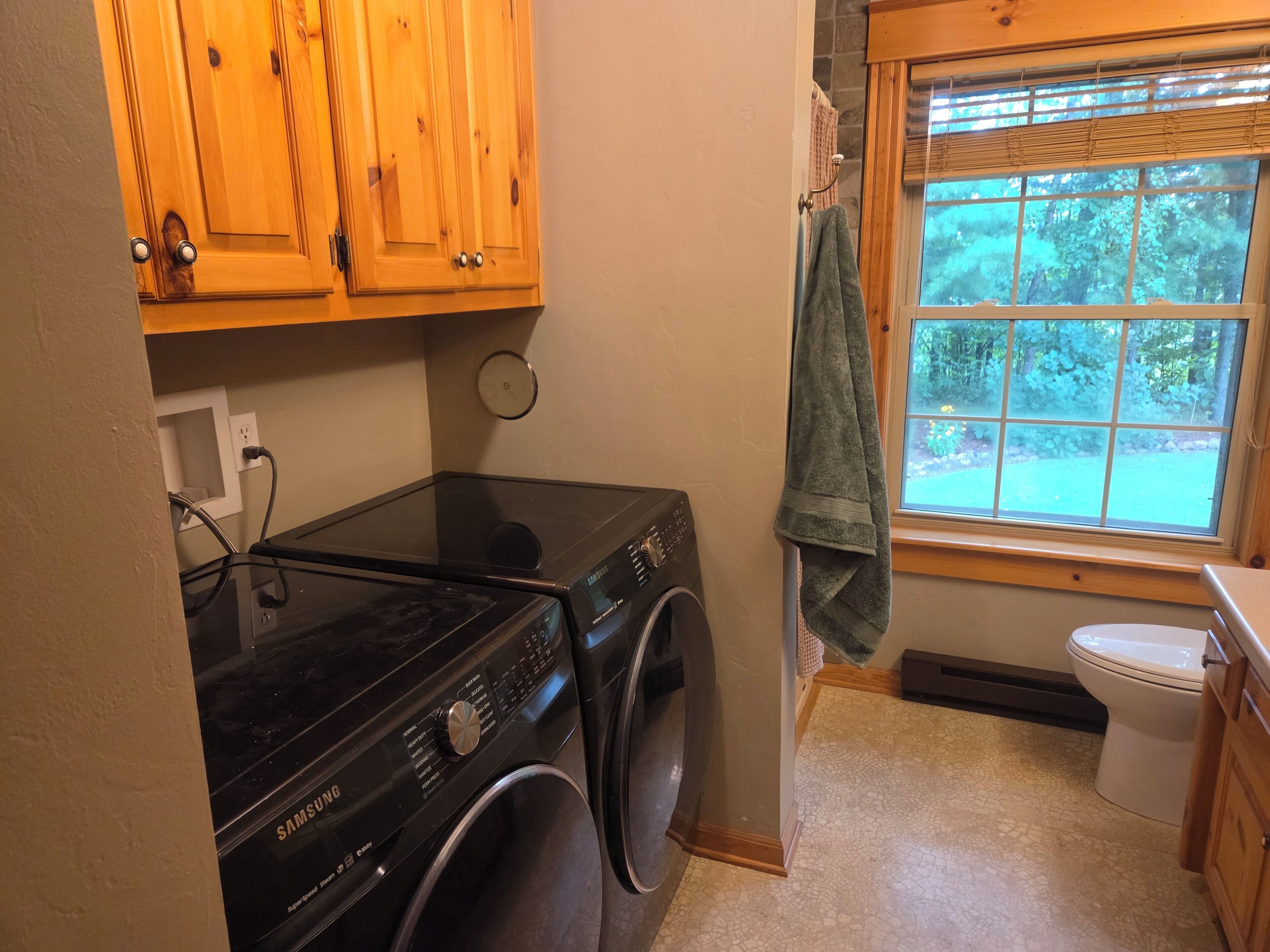 A laundry room with a Samsung washer and dryer, wooden cabinets, a window with a view of greenery, and a toilet.