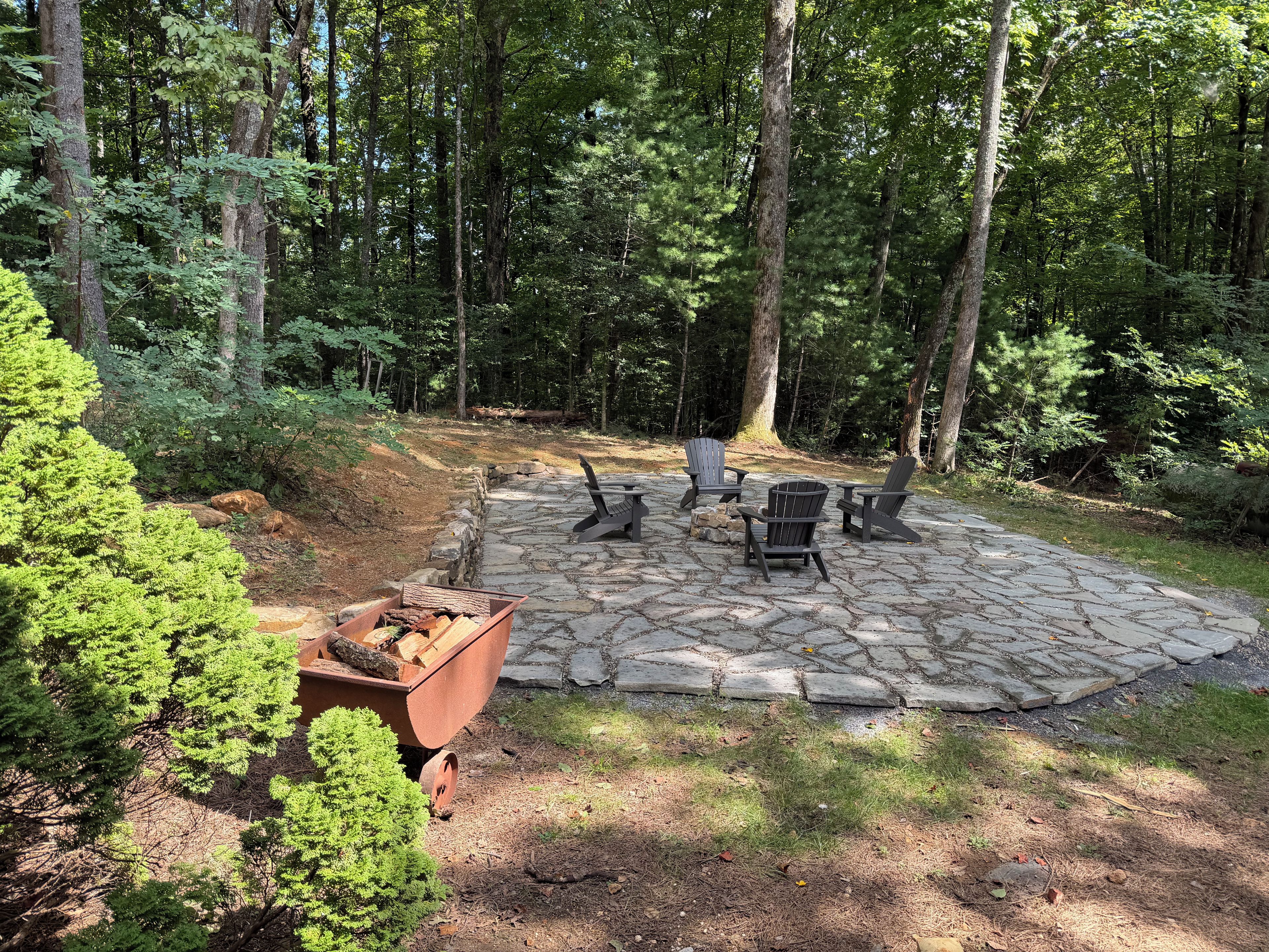 A stone patio with Adirondack chairs and a fire pit surrounded by a wooded area.