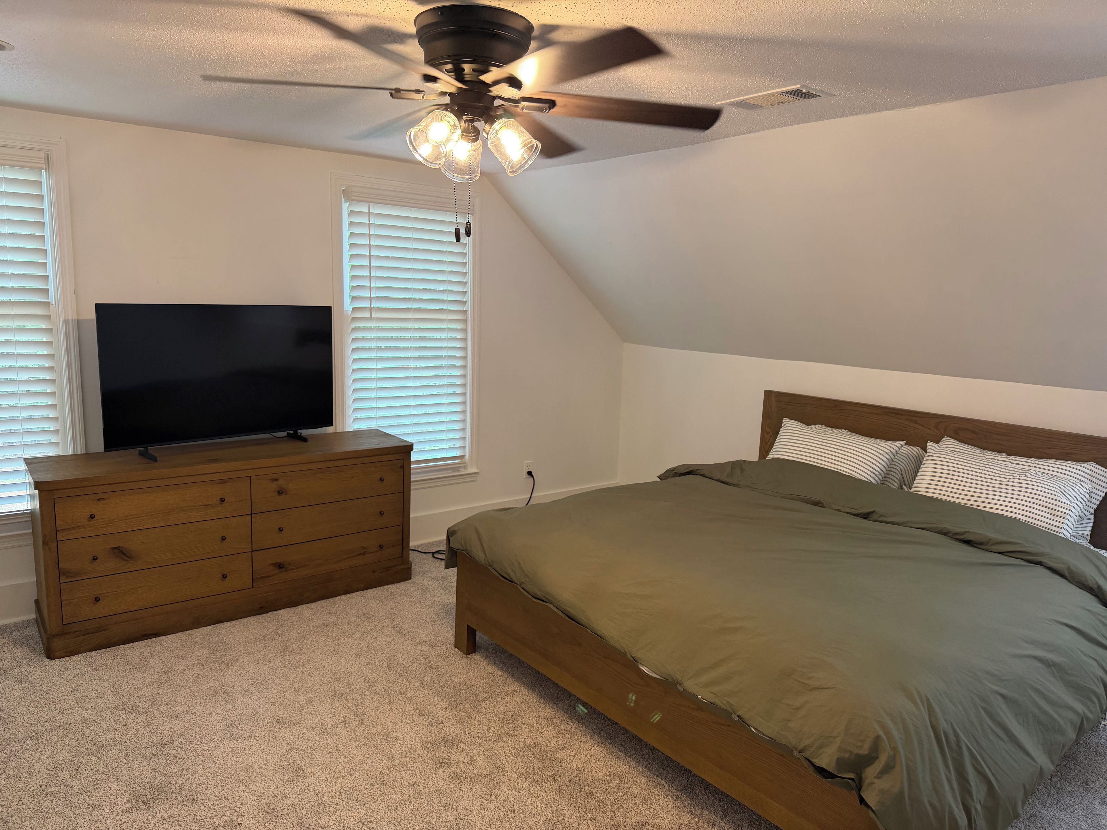 A bedroom with a bed, dresser, TV, ceiling fan, and windows.