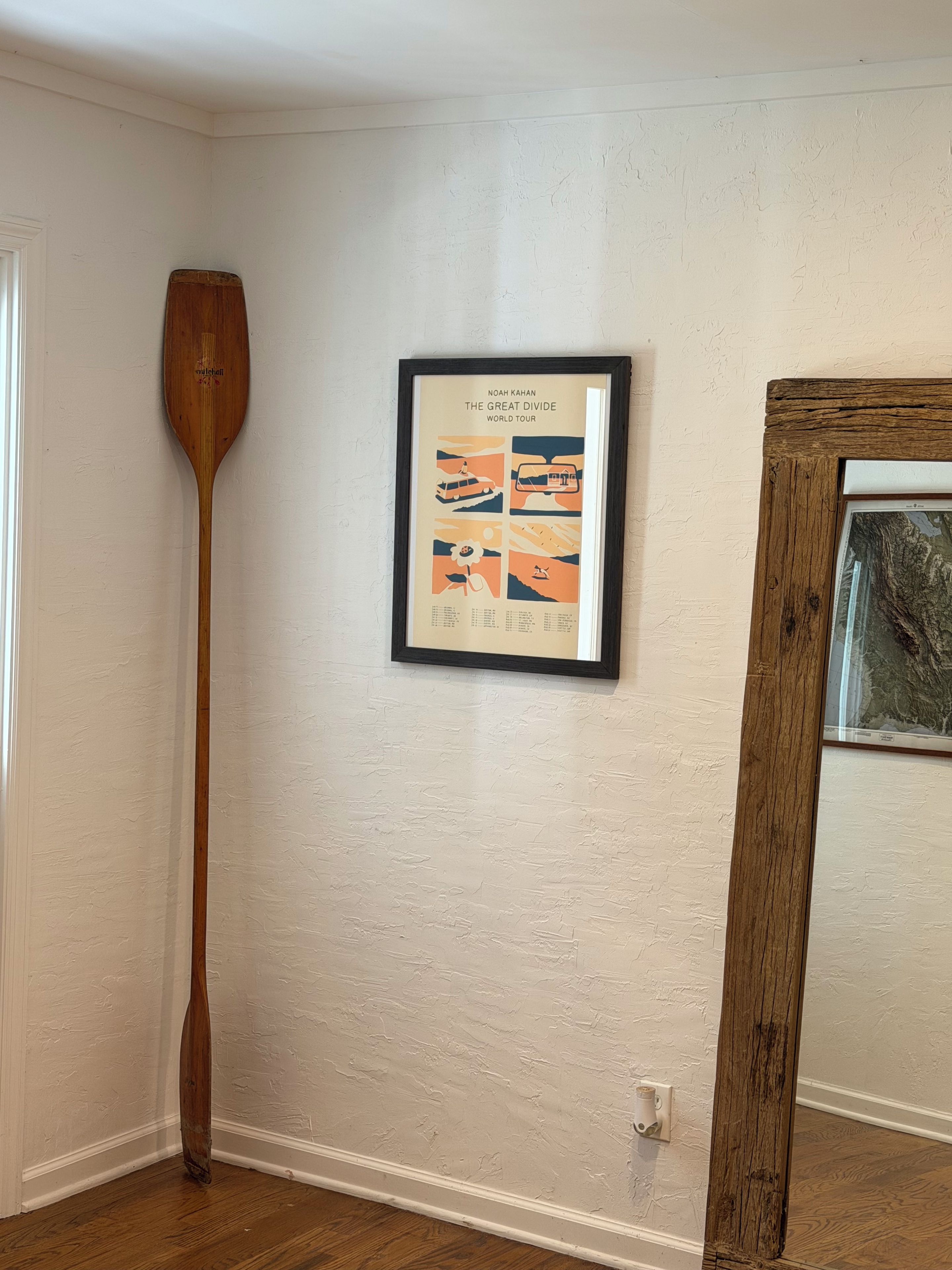 Ein gerahmtes Poster an einer Wand, ein hölzernes Paddel, das an die Wand gelehnt ist, und ein gerahmtes Bild zur Rechten.