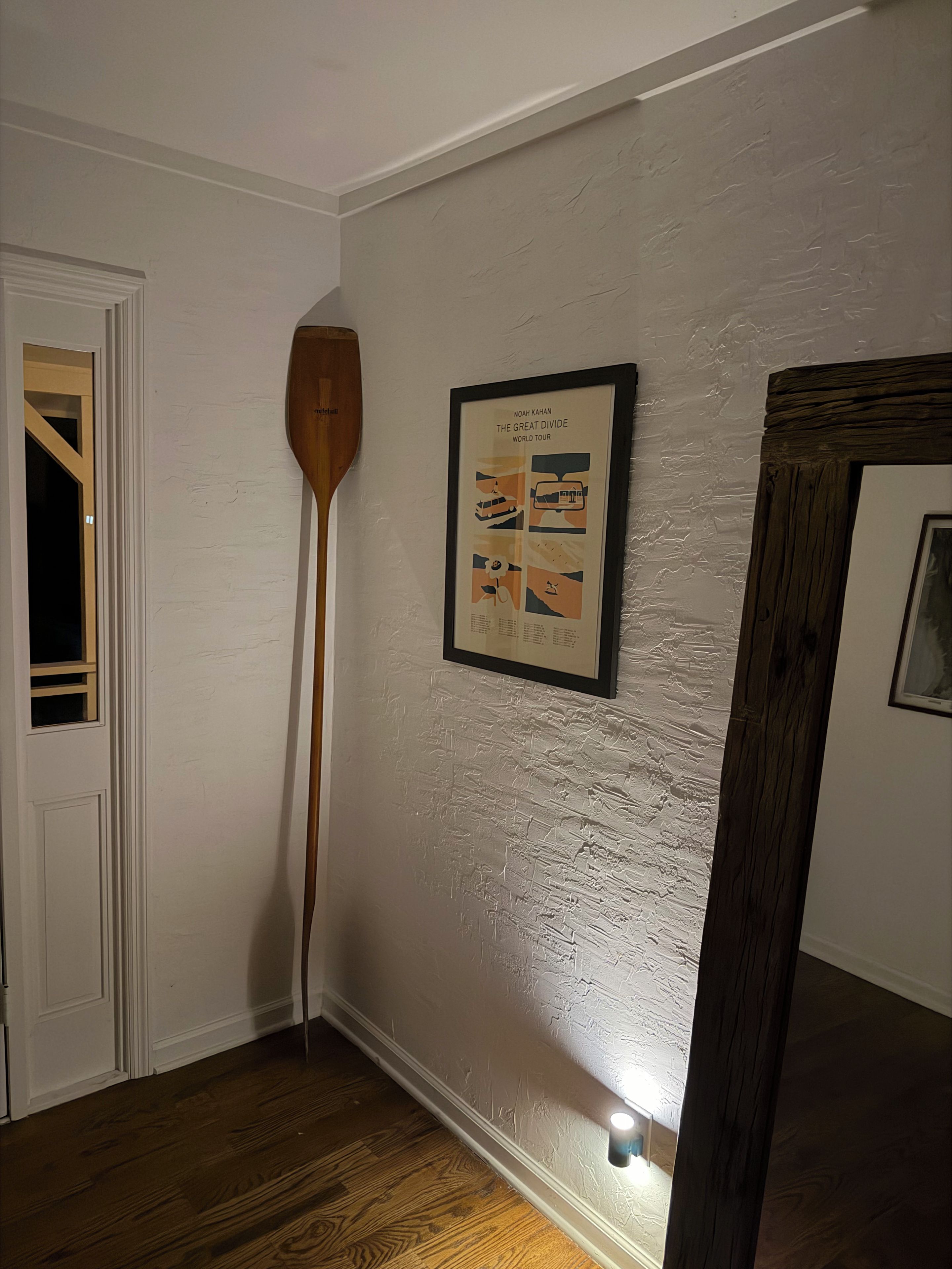 A room with a framed poster on the wall, a wooden paddle leaning against the wall, and a door with a window.
