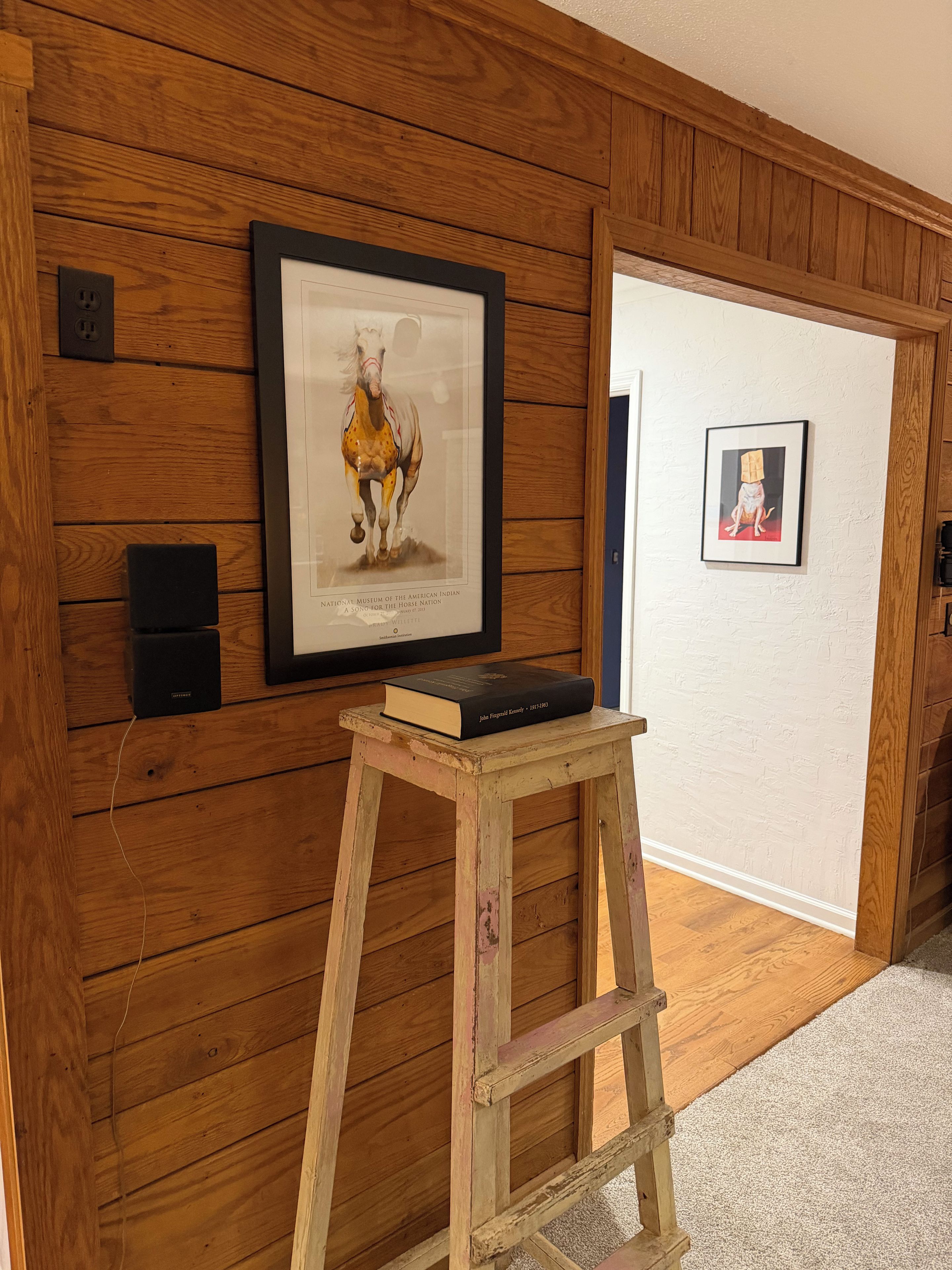 A wooden wall with framed artwork, a speaker, and a ladder with a book.