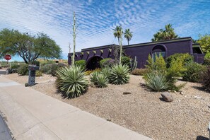 Un bâtiment à la façade violette, un panneau de signalisation de type « stop », et une variété de plantes désertiques.