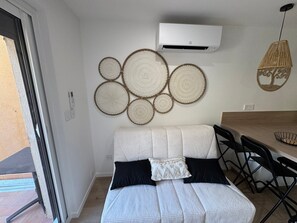 A room with a bed, a dining table with chairs, a wall-mounted air conditioner, and a woven wall decoration. - Le Petit Coin Bohéme, 4 pers, pieds dans l'eau (Le Cap d'Agde)