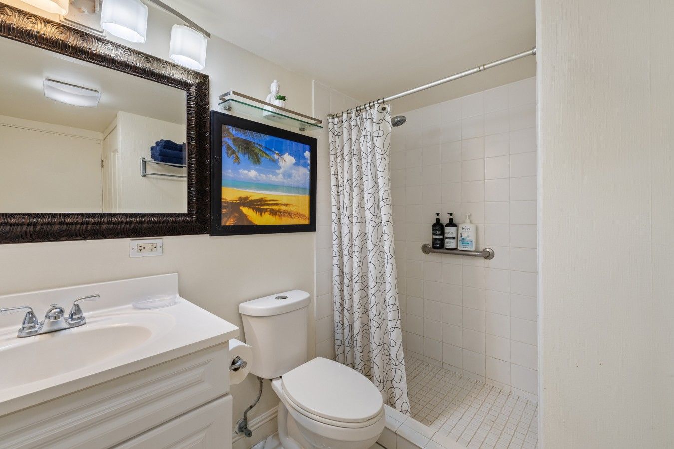 Ein Badezimmer mit einem weißen Waschbecken, einer Toilette und einer Duschvorhang. Über dem Waschbecken hängt ein gerahmtes Bild, und eine Ablagefläche bietet Platz für Toilettenartikel.