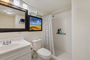 Ein Badezimmer mit einem weißen Waschbecken, einer Toilette und einer Duschvorhang. Über dem Waschbecken hängt ein gerahmtes Bild, und eine Ablagefläche bietet Platz für Toilettenartikel.