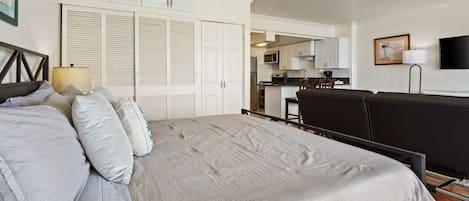A bedroom with a bed, pillows, a bedside table, a lamp, a TV, and a kitchenette in the background.