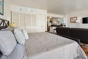 Une chambre à coucher avec un lit, des oreillers, une table de chevet, une lampe, un téléviseur et, au fond, une petite cuisine.
