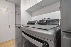 A laundry room with a front-loading washer and dryer, white cabinets, and a wooden floor. - Two-home tropical retreat near Palm Beach Zoo (West Palm Beach)