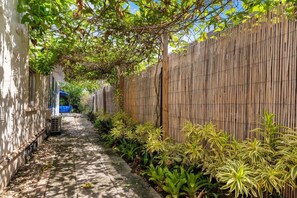 A narrow pathway flanked by a bamboo fence and lush greenery, leading to a building with a blue awning. - Two-home tropical retreat near Palm Beach Zoo (West Palm Beach)
