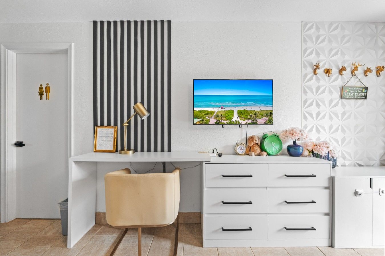A modern room with a white desk, a chair, a television displaying a beach scene, and a decorative wall with geometric patterns.
