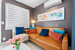 Een woonkamer met een bruine leren bank, blauwe en rode kussens, een witte bijzettafel, een lamp en een vaas met roze bloemen.