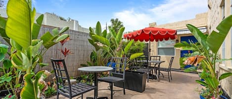Un patio al aire libre con mesas y sillas, rodeado de plantas en macetas y un paraguas rojo.
