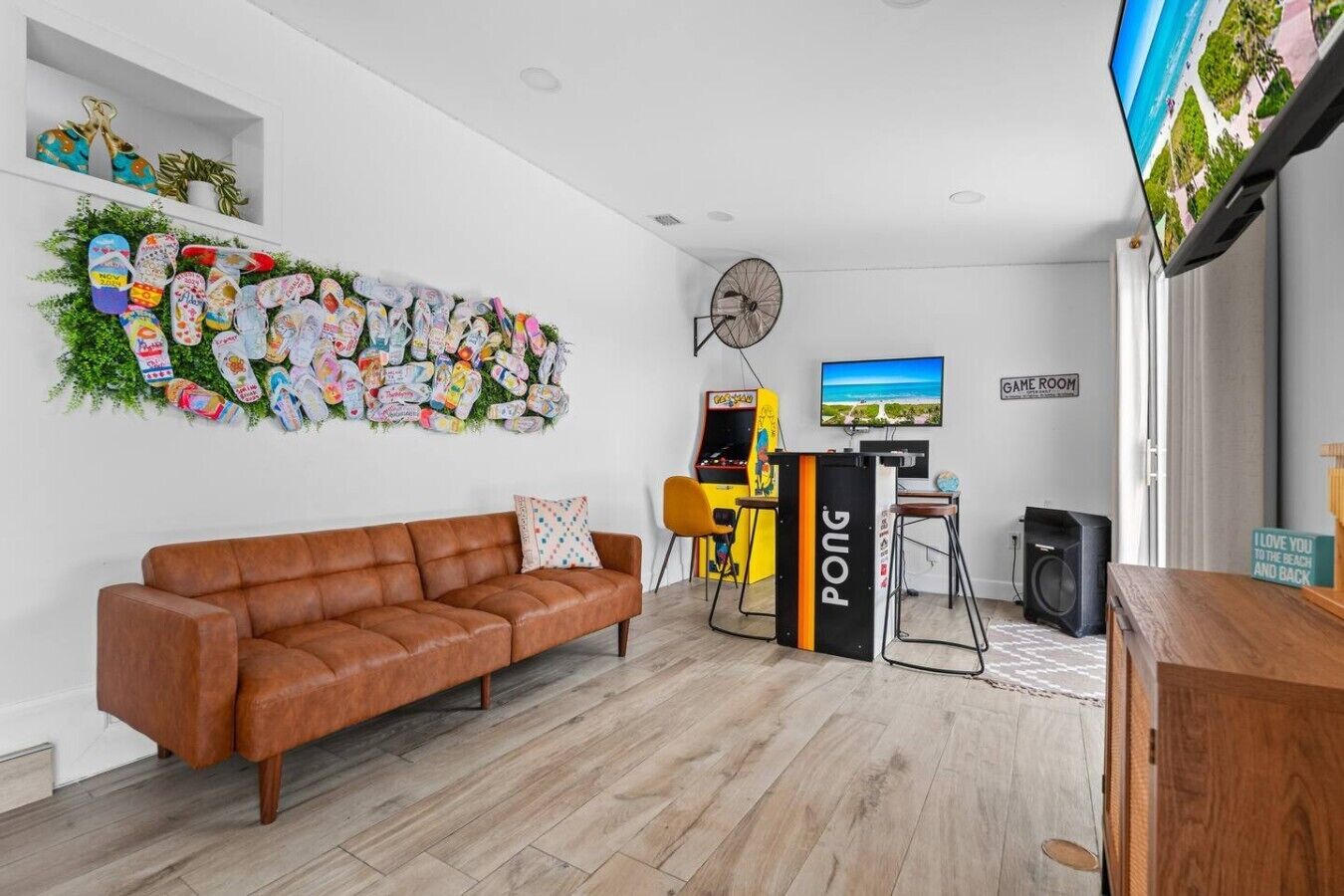 Una sala de estar con un sofá de cuero marrón, una pared decorada con diversos objetos, una consola de videojuegos y un televisor que muestra una escena de playa.