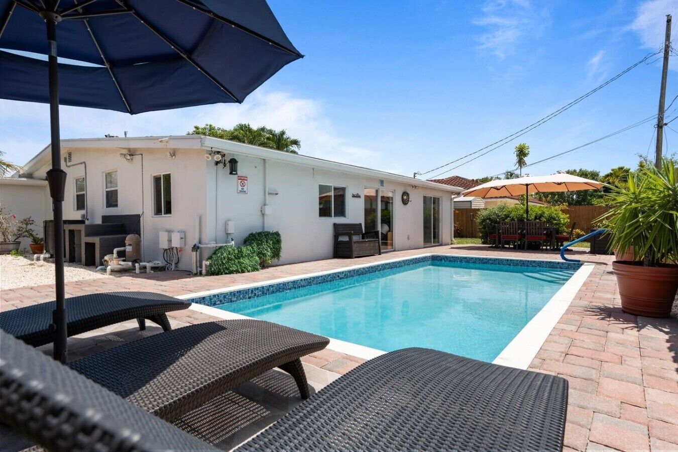 Une piscine avec des chaises longues et des parasols, un bâtiment blanc doté d’une porte et de fenêtres, et un espace clos agrémenté de plantes.
