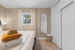 Ein Hotelzimmer mit einem Bett, einem Nachttisch mit Lampe, einem Fenster mit Vorhängen, einem Spiegel und einem kleinen Hocker.