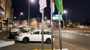 Una calle nocturna con fila de coches aparcados, un edificio blanco y tres banderas en mástiles.