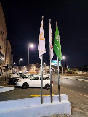 Una calle nocturna con fila de coches aparcados, un edificio blanco y tres banderas en mástiles.
