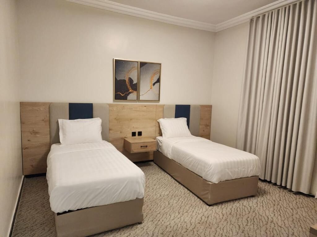 Ein Hotelzimmer mit zwei Einzelbetten, einem Holzkopfteil, einem Nachttisch und einem großen Fenster mit Vorhängen.
