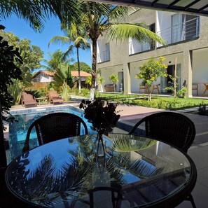Ein Tisch mit Glasplatte, Korbstühle, ein Pool und eine Terrasse mit Blick auf ein modernes Gebäude und Palmen.