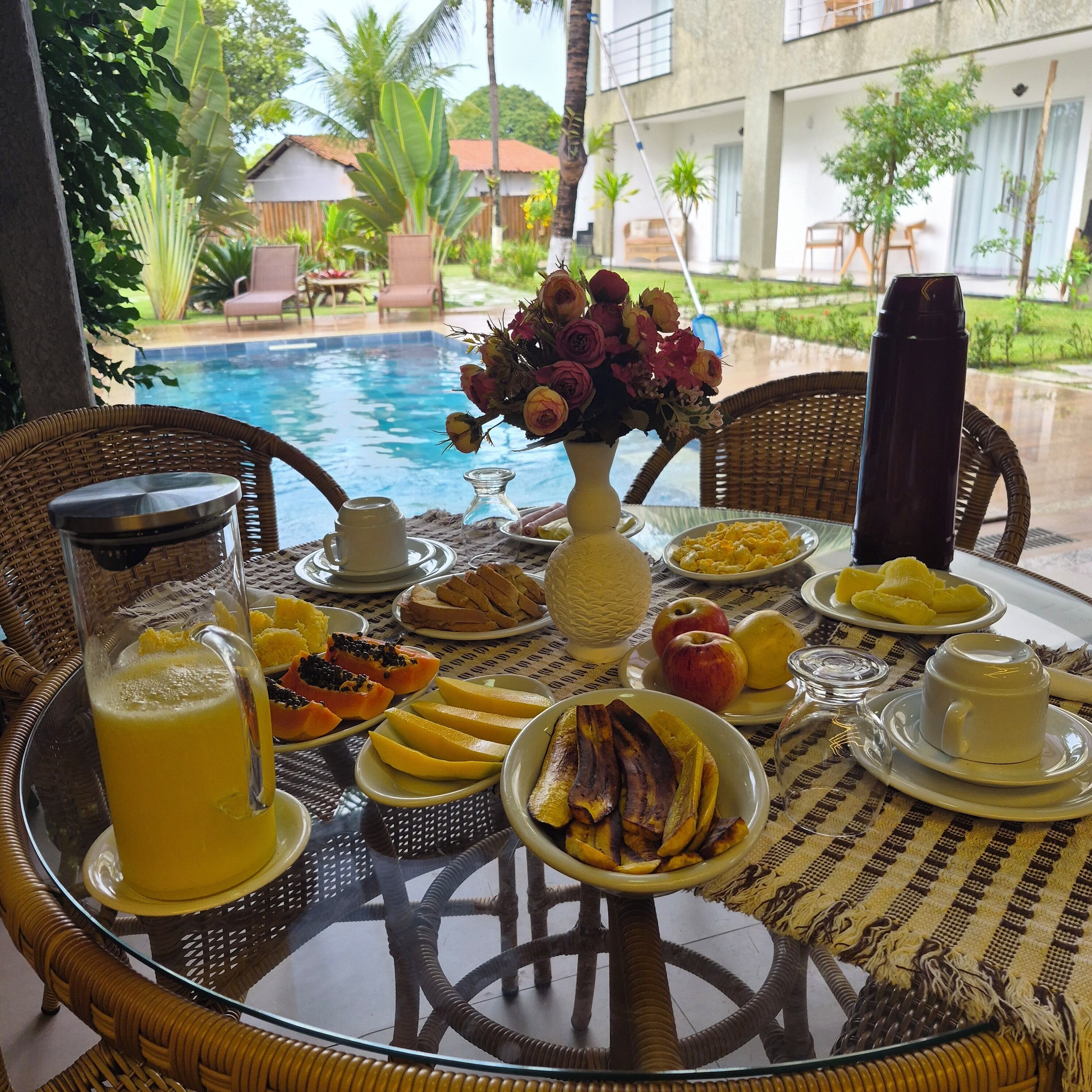 Une table à manger avec des aliments pour le petit-déjeuner, un vase de fleurs et une vue sur une piscine et un jardin.
