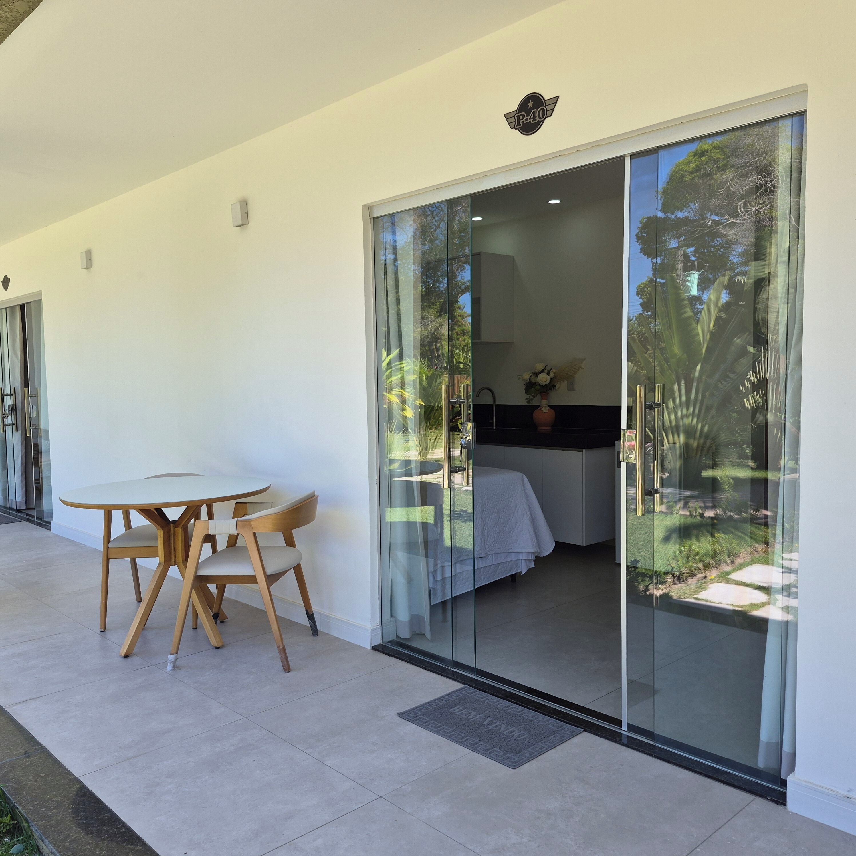 Quarto moderno com porta de vidro deslizante para um jardim, uma pequena mesa redonda com cadeiras e uma cama com cobertura branca.