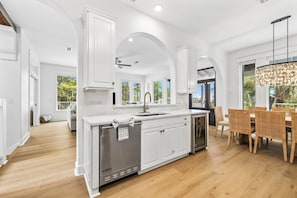 A modern kitchen with white cabinetry, a stainless steel dishwasher, and a dining area with wicker chairs. - Close to Beach | Private Pool | Games | LSV (Santa Rosa Beach)