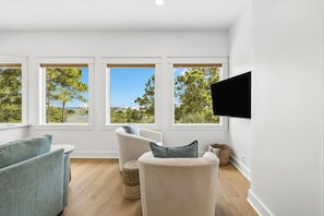 Ein Zimmer mit an der Wand montiertem Fernseher, drei Fenstern mit Blick auf Bäume und eine entfernte Landschaft, ein beiges Sofa und ein Korbwäschekorb.