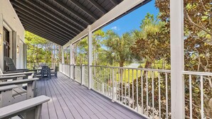 Eine überdachte Terrasse im Freien mit Grill und Stühlen, umgeben von Bäumen und wolkenlosem Himmel.