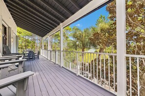 Eine überdachte Terrasse im Freien mit Grill und Stühlen, umgeben von Bäumen und wolkenlosem Himmel.
