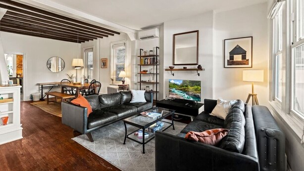 A modern living room with a black leather sofa, a coffee table, and a TV. There is a bookshelf and a dining area with a table and chairs. - Charming 3BR East Falls Home with Fenced Yard & Office (Philadelphia)