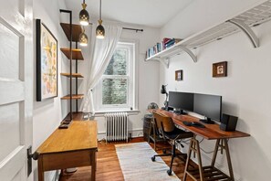 Ein Zimmer mit einem Holztisch, einem Computer, einem Stuhl, einer Heizung, einem Fenster mit Vorhängen und einem Bücherregal.