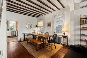 Uma área de refeições com uma mesa de madeira, cadeiras e um banco. Há um radiador sob uma janela, um abajur sobre uma mesinha e uma estante à direita.