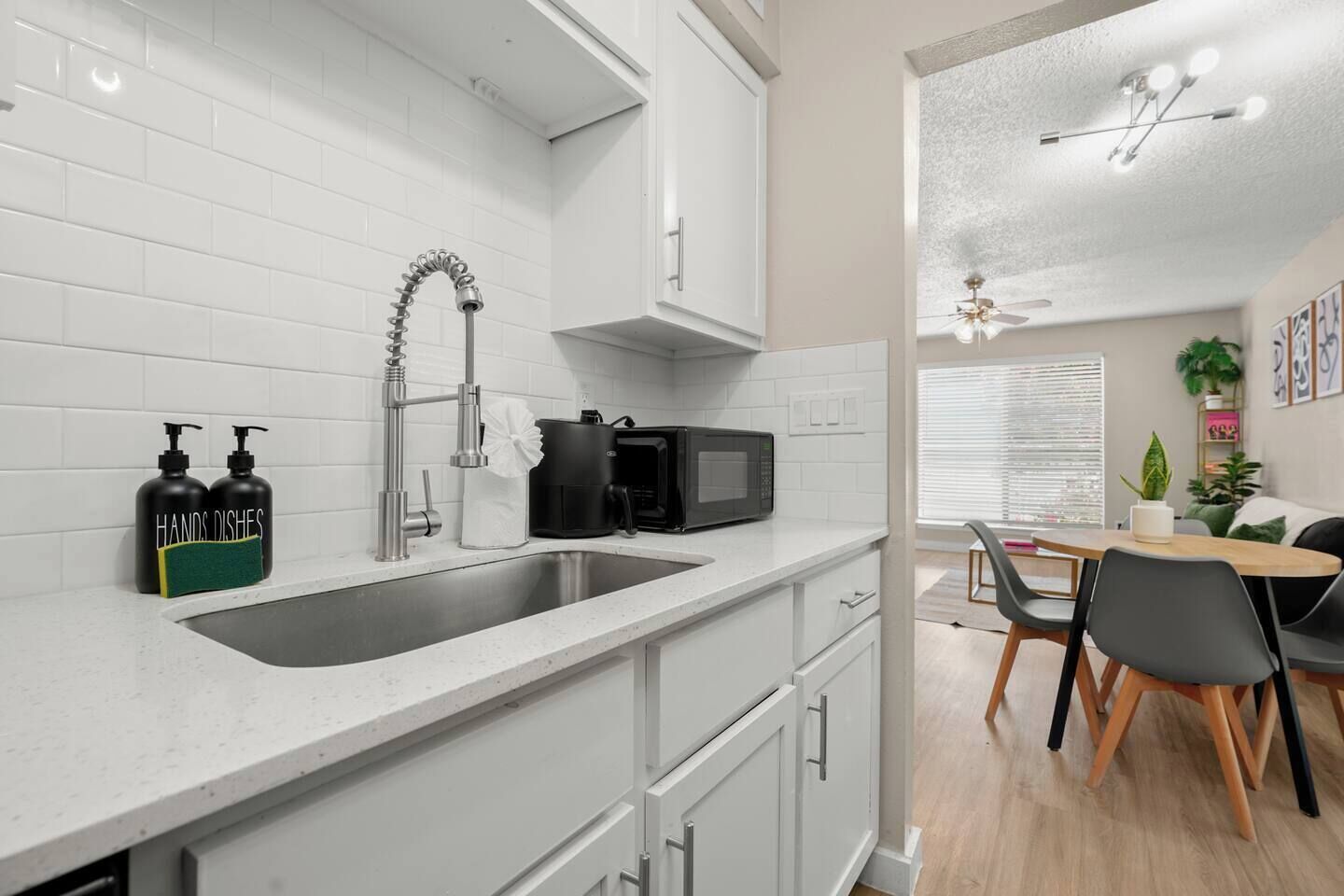 A modern kitchen with a stainless steel sink, a coffee maker, and a microwave. There is a dining area with a table and chairs.