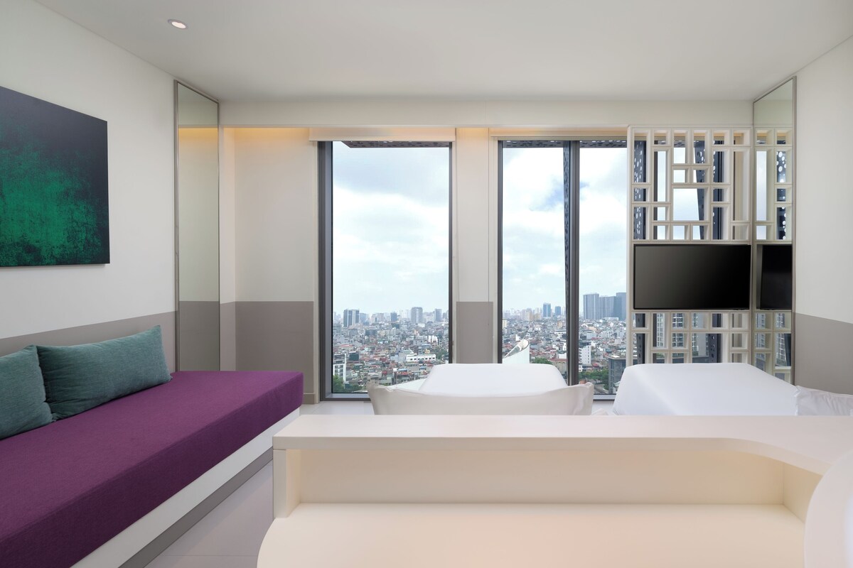 A modern hotel room with a large window, a bed, a sofa, and a TV.