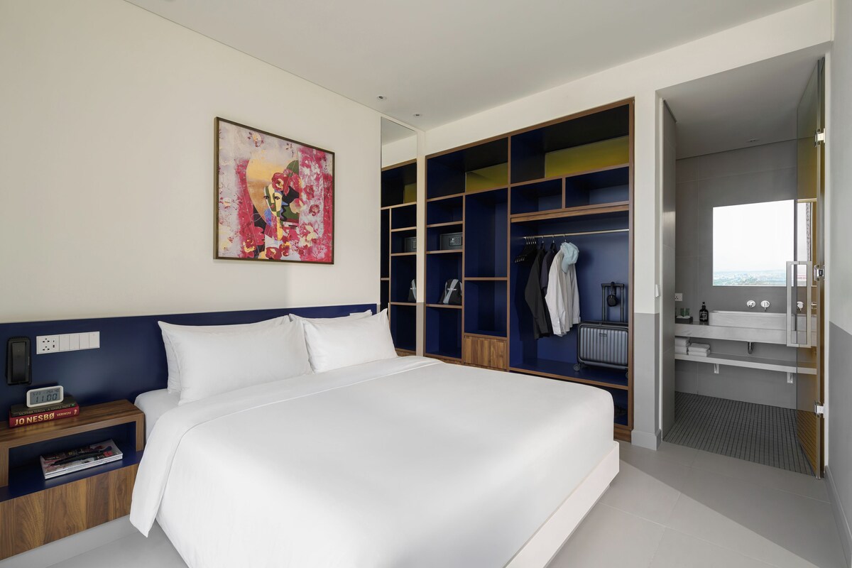 A modern hotel room with a large bed, a wardrobe with multiple compartments, a bathroom with a vanity, and a painting on the wall.