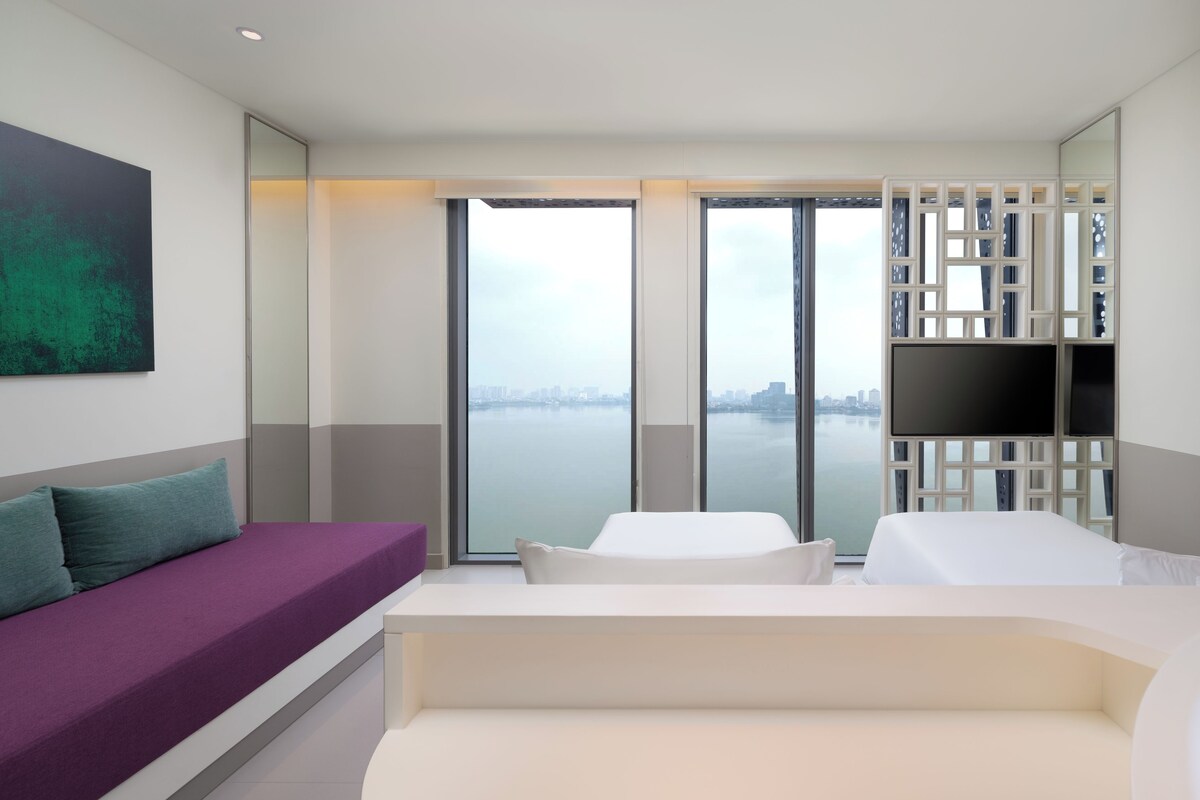 A modern hotel room with a large window, a sofa, a bed, and a TV.