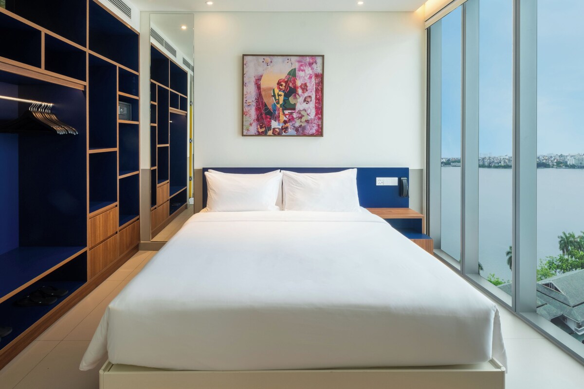 A hotel room with a large bed, a wardrobe with multiple compartments, a painting on the wall, and a view of the city through the window.