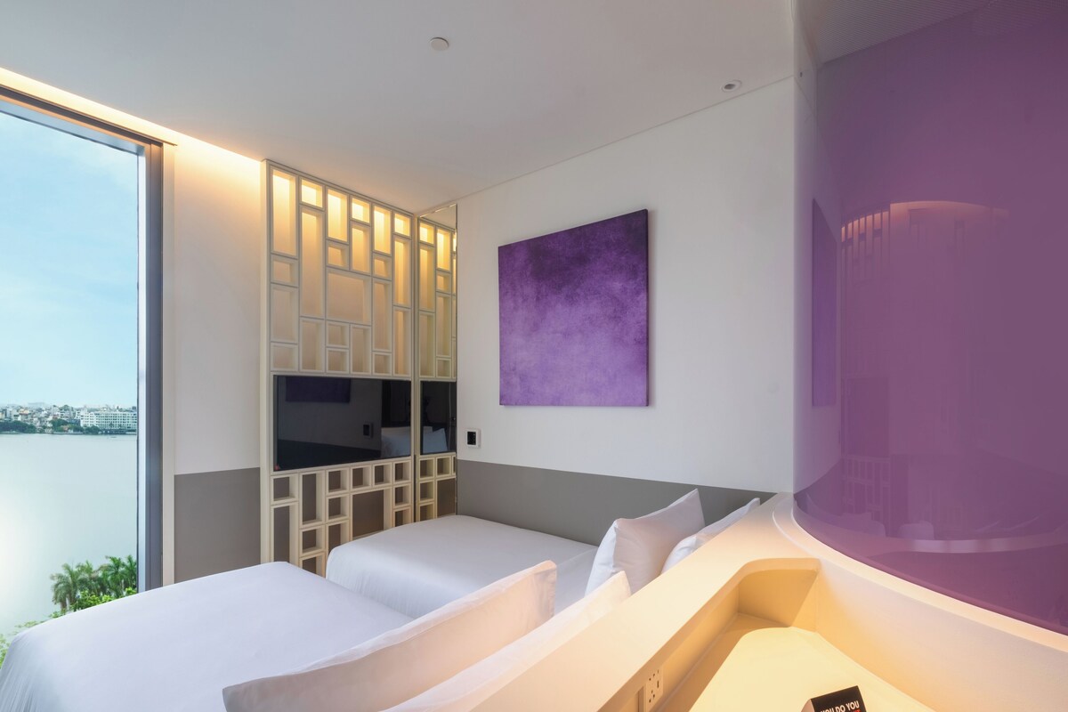 A hotel room with two beds, a large window, a purple artwork, and a view of the city.