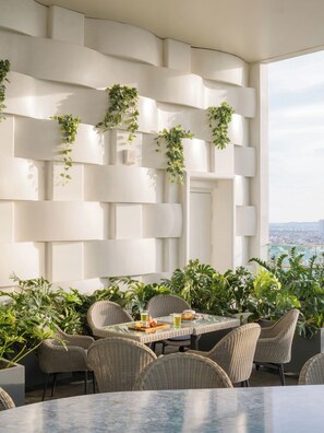 A modern outdoor seating area with wicker chairs and a table, surrounded by lush greenery and a white wall with geometric patterns. - Wink Hanoi Westlake, Unscripted by Hyatt - 24 Hours stay (Hanoi)