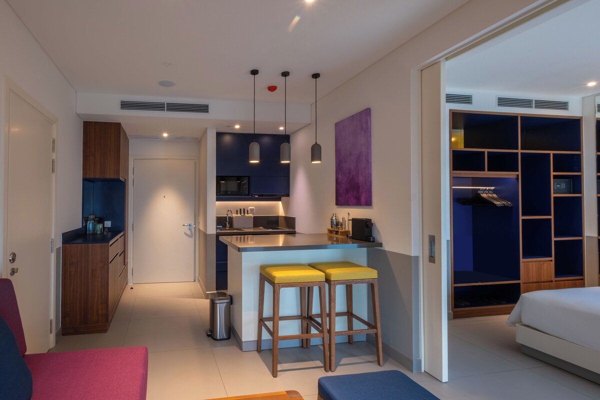 A compact hotel room with a kitchenette, a bed, a seating area with stools, and a wardrobe with built-in shelving.