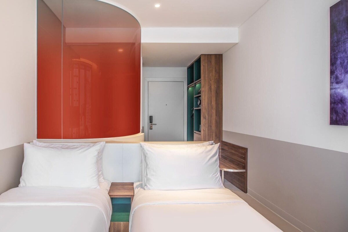 A hotel room with two beds, a wooden headboard, a mirror, a wardrobe, and a painting on the wall.