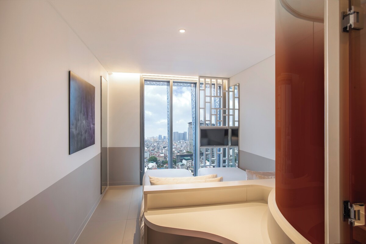 A modern hotel room with a city view, a bed, a desk, and a large window with a view of tall buildings.