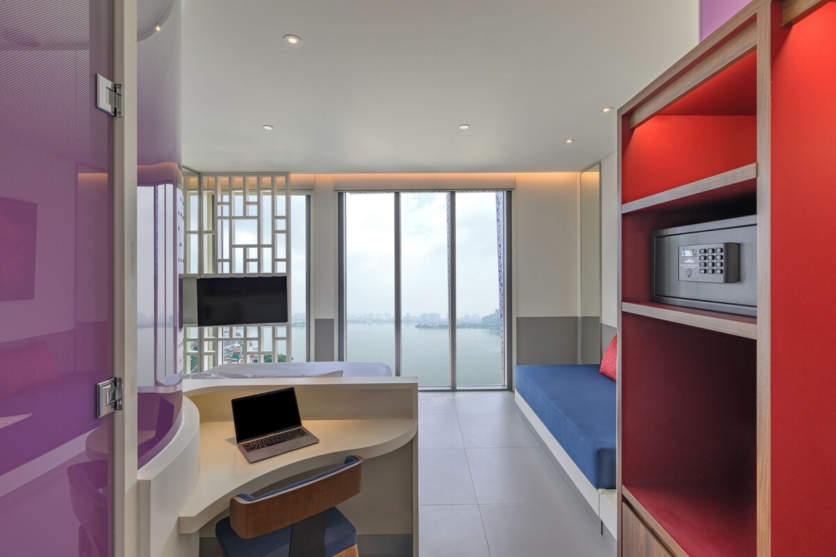 A modern hotel room with a laptop on a desk, a bed with a blue headboard, a red shelving unit, and a view of a cityscape through large windows.