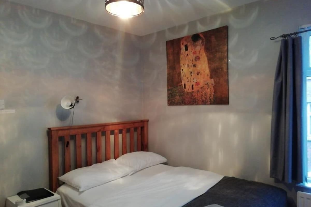 A bedroom with a wooden bed, a bedside table, a wall-mounted lamp, and a painting of a castle.