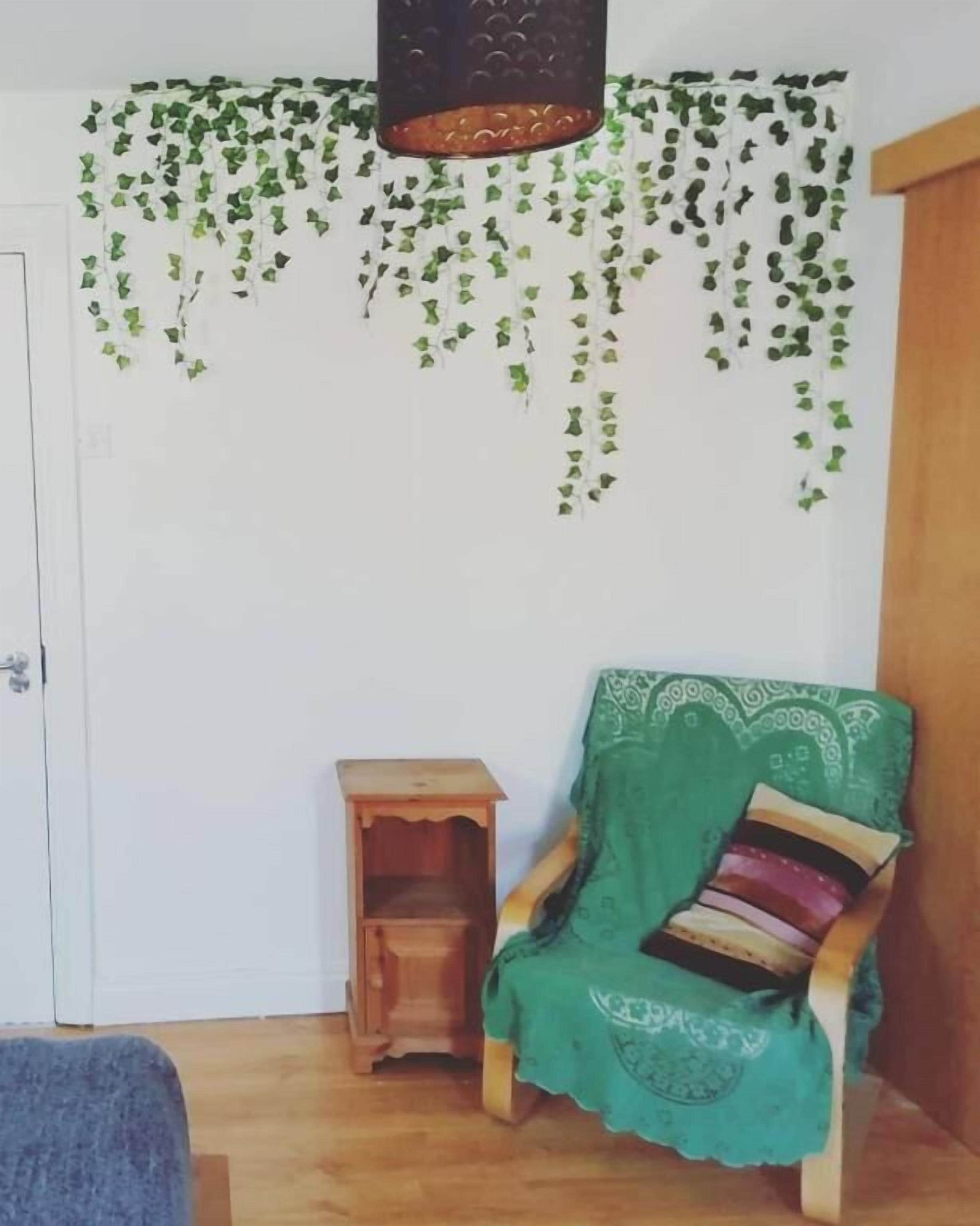 A room with a wooden floor, a green chair with a patterned cushion, a small wooden side table, and a hanging plant with green leaves.