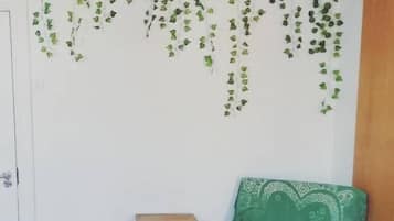 A room with a wooden floor, a green chair with a patterned cushion, a small wooden side table, and a hanging plant with green leaves.