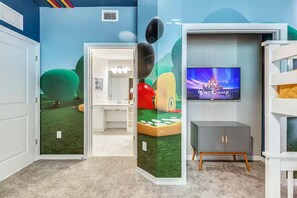 Ein Zimmer mit einer Wandmalerei einer grünen Landschaft, einem Fernseher der das Walt Disney Logo zeigt und einem grauen Schrank mit Holzbeinen.