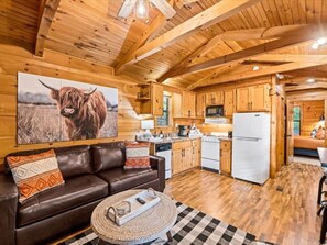 Ein Holzblockhaus-Interieur mit Küche und Wohnbereich, ausgestattet mit einer Couch, einem Esstisch und einem großen Gemälde einer Kuh.